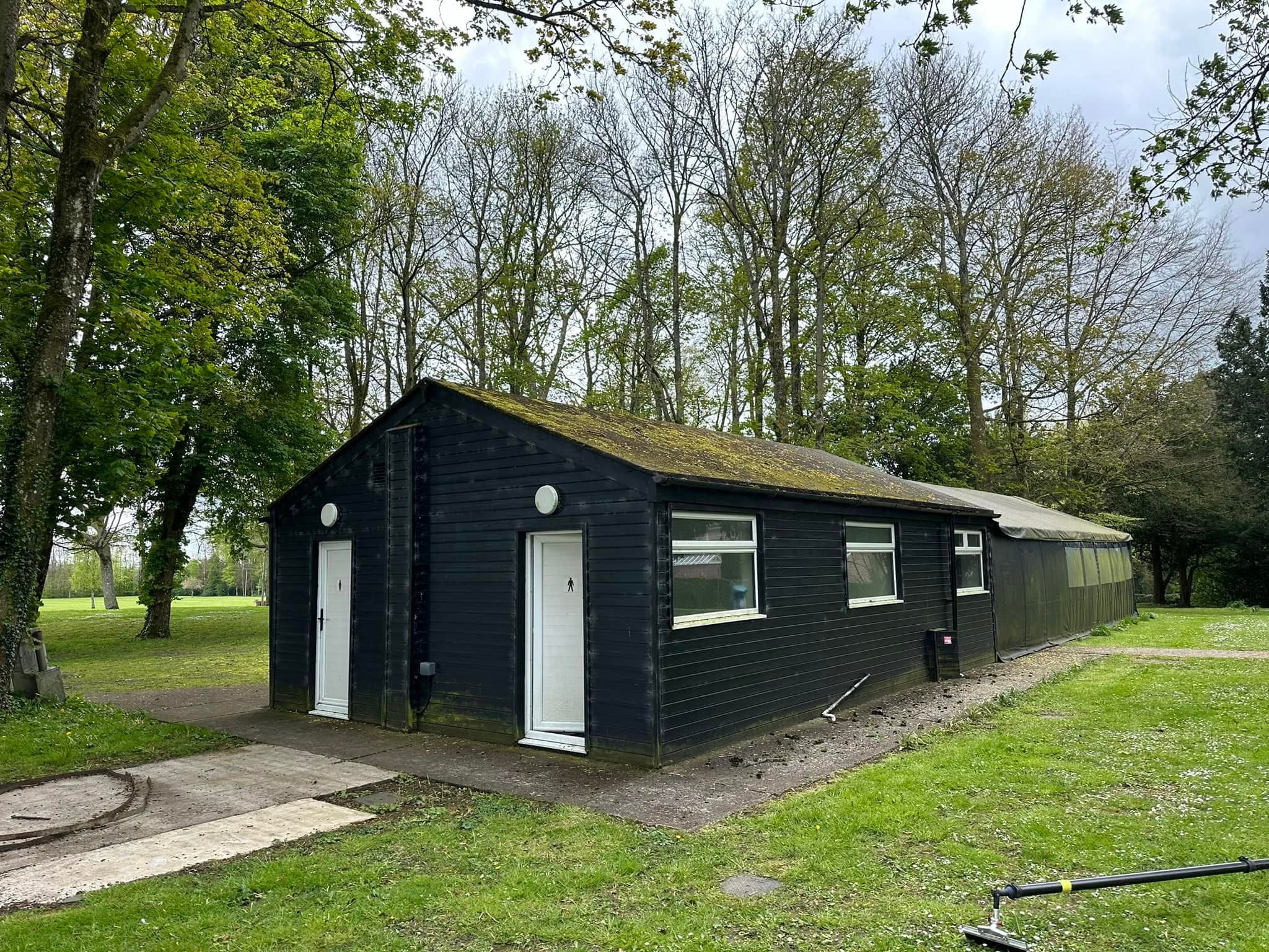 Long black wooden building with white doors and windows in a grassy park with trees.