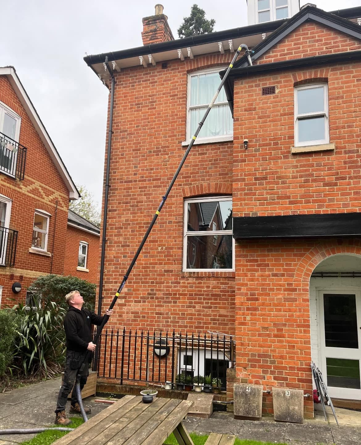 Man cleaning gutters on a two-story red brick house using a long telescopic pole.