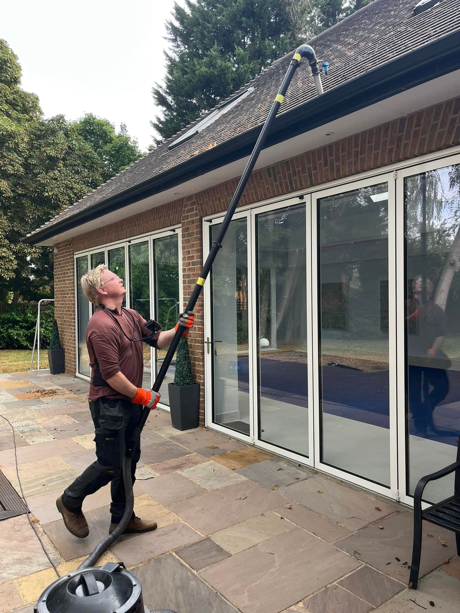 Man using a long telescopic vacuum pole to clean gutters on a brick house.