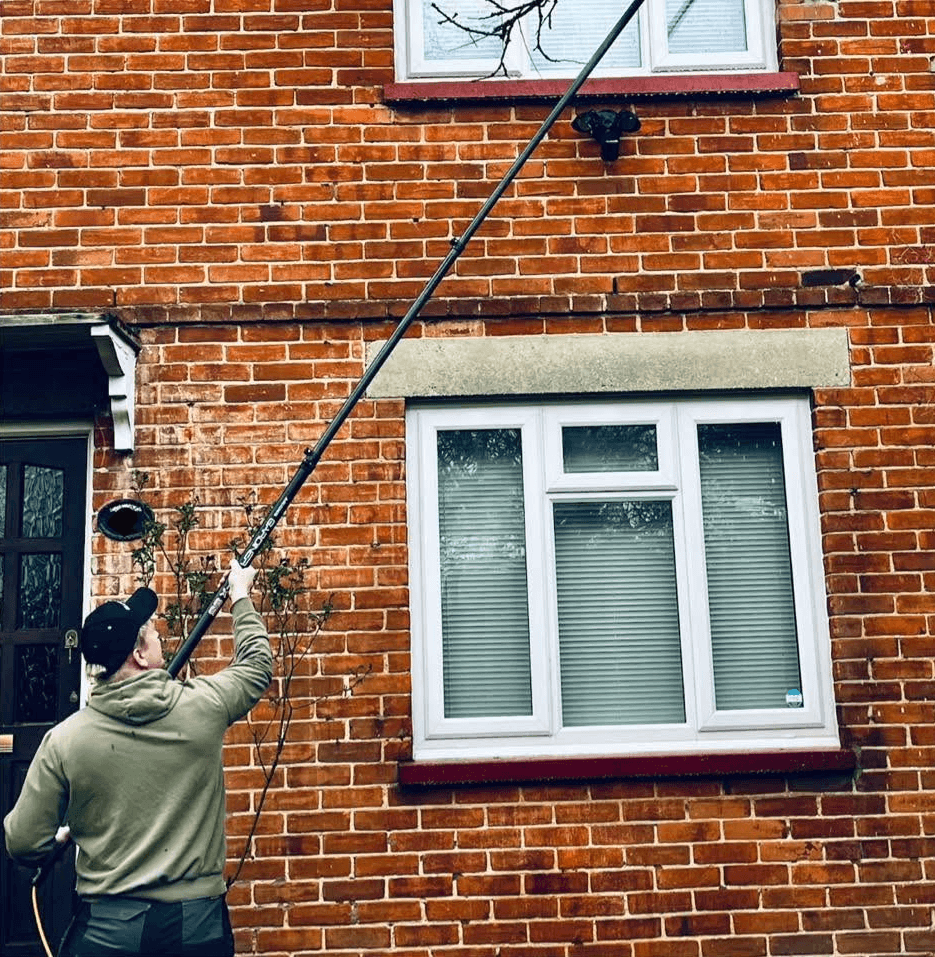 Man using a long telescopic pole to clean upper-story windows of a red brick house.