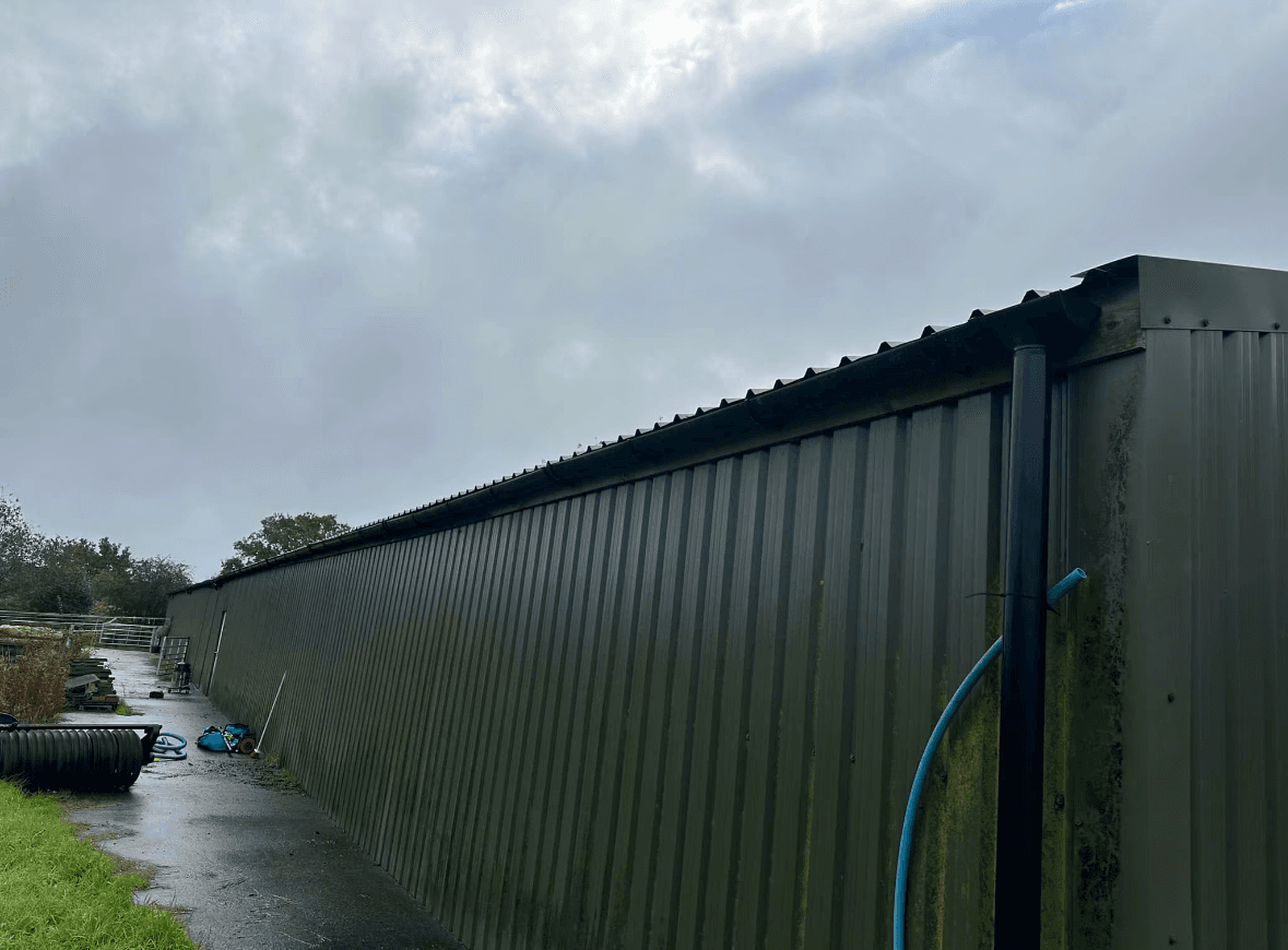 Long green corrugated metal building with black gutters and downpipe under a cloudy, overcast sky.