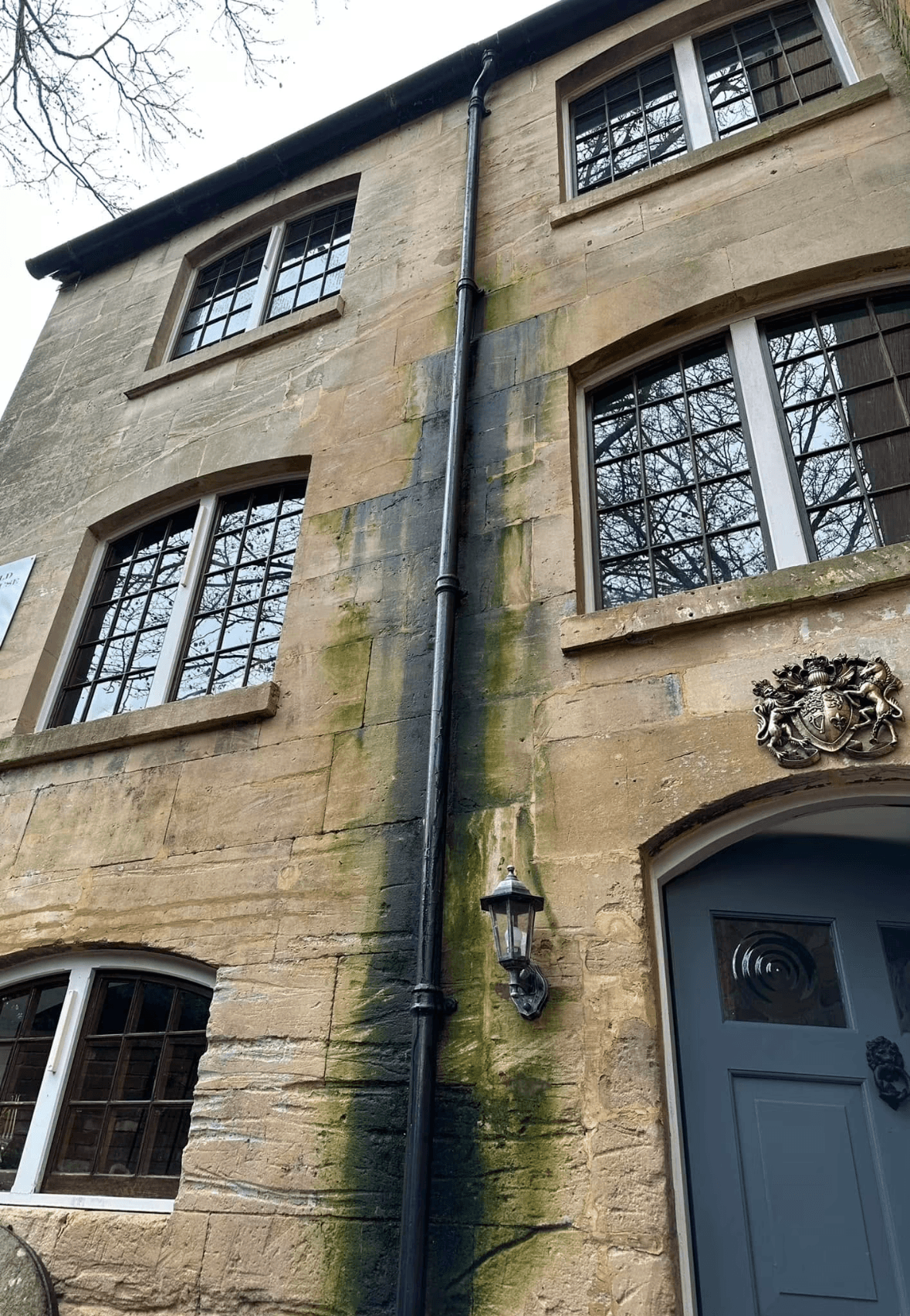 Green algae stains a stone building wall around a leaking black vertical drainpipe.