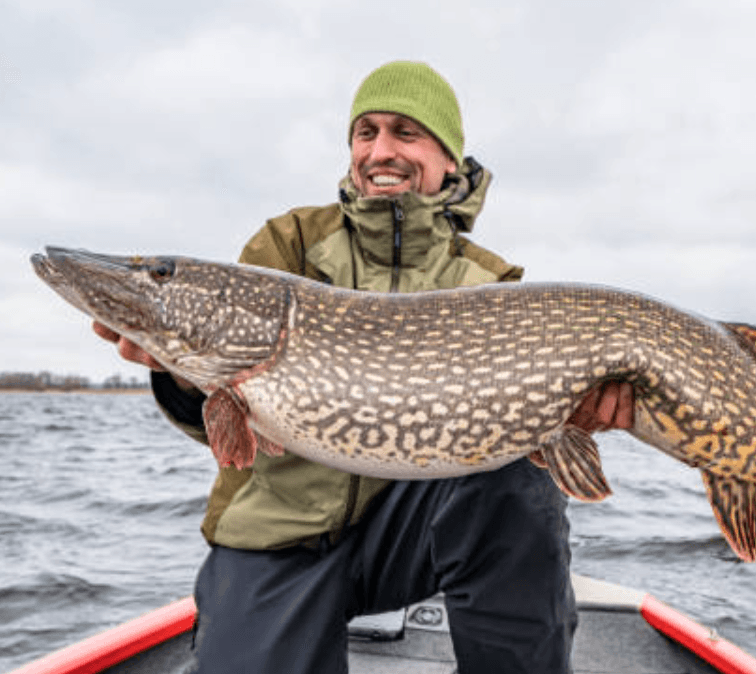 Smiling fisherman in a green beanie holding a large northern pike on a boat.